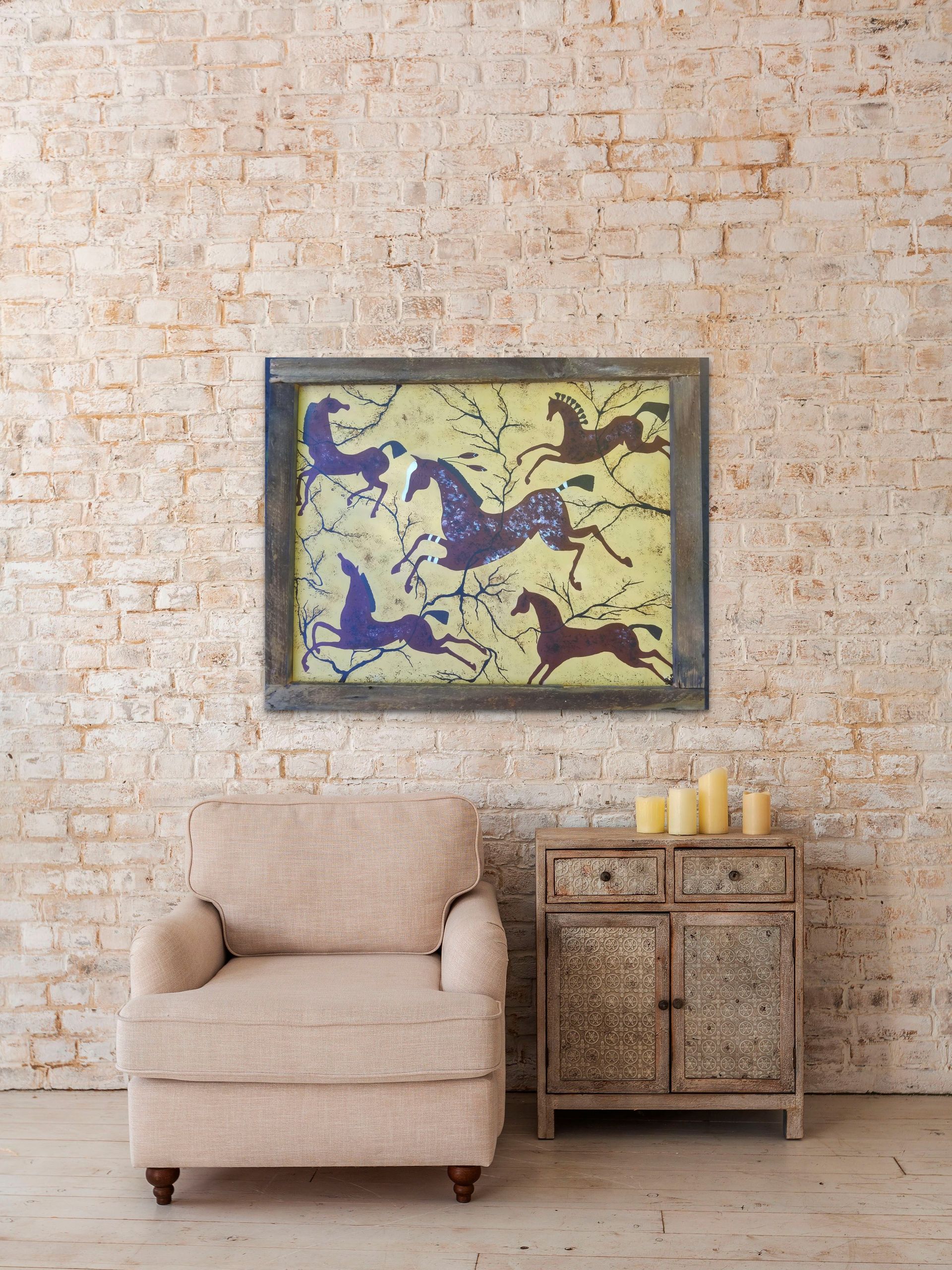 A beige armchair and rustic cabinet sit before a brick wall displaying a framed print of horses on a light yellow ground.