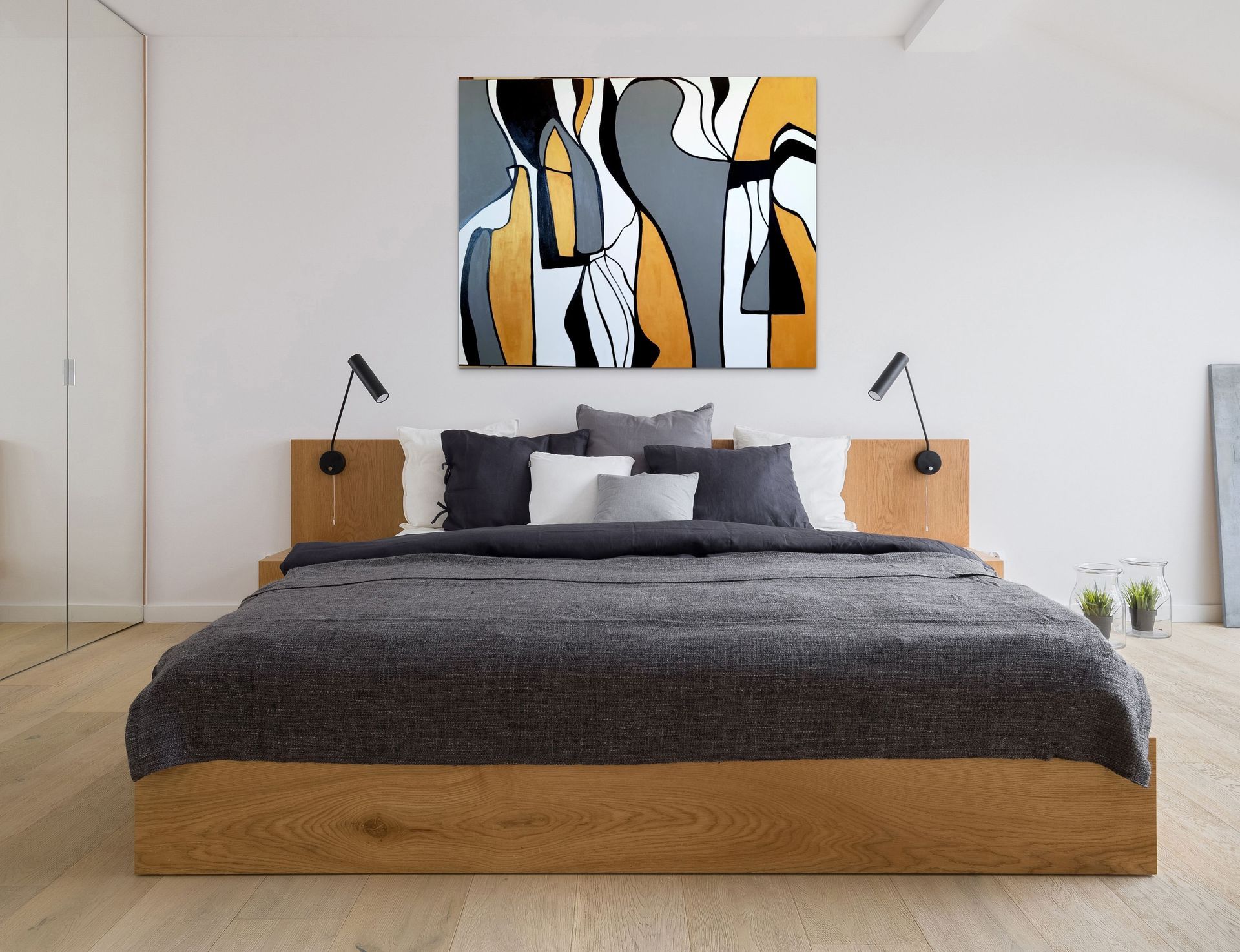 A modern bedroom with a low wood-frame bed, dark gray bedding, wall-mounted reading lights, and abstract wall art.