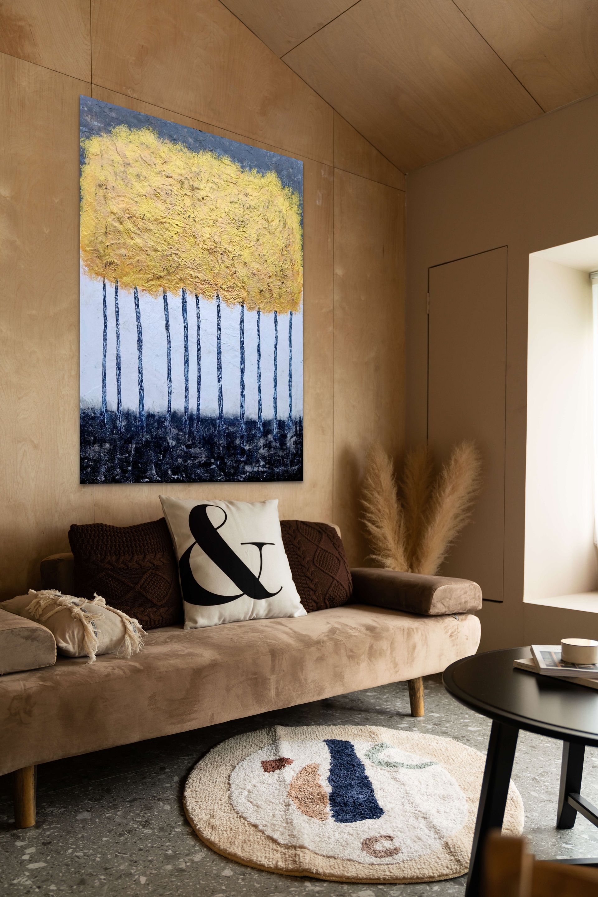 A modern living room features a large, vertical painting of gold-leafed trees hanging above a neutral sofa with pillows.