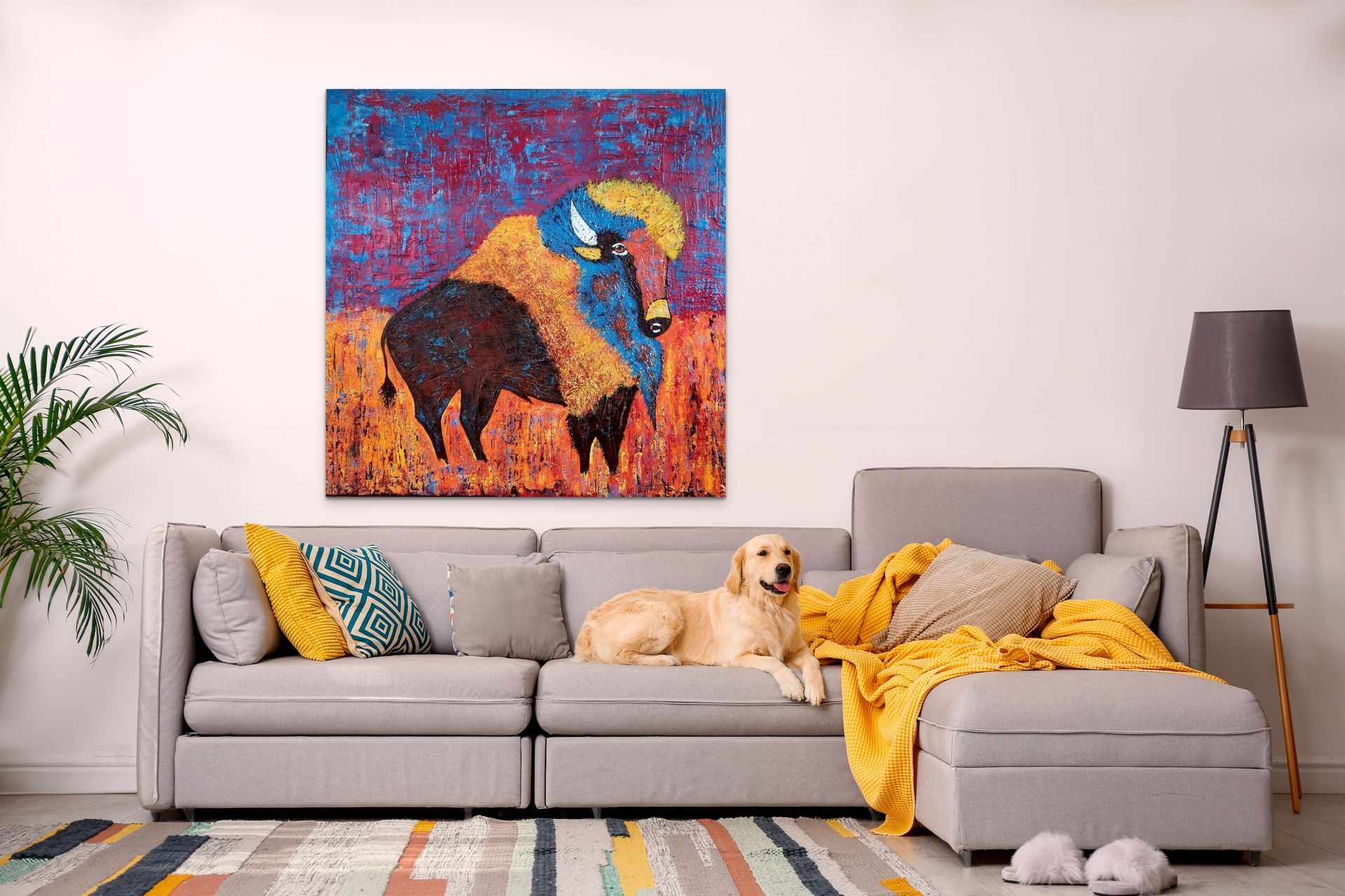 A light-colored dog sits on a gray sectional sofa beneath a large, colorful painting of a buffalo in a modern living room.