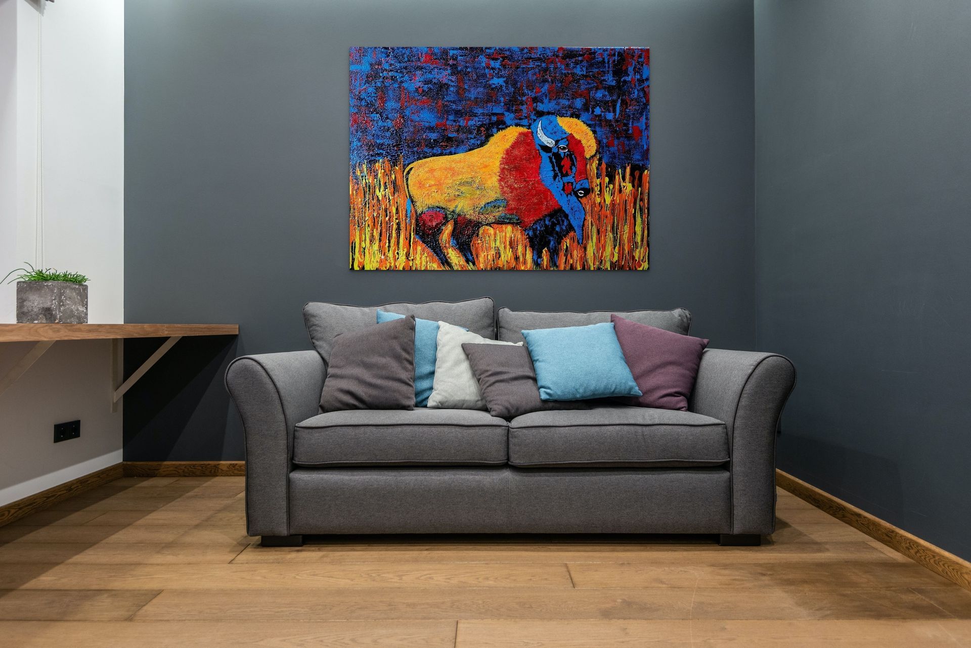 A grey sofa with colorful pillows sits against a dark wall under a vibrant abstract painting of a buffalo.