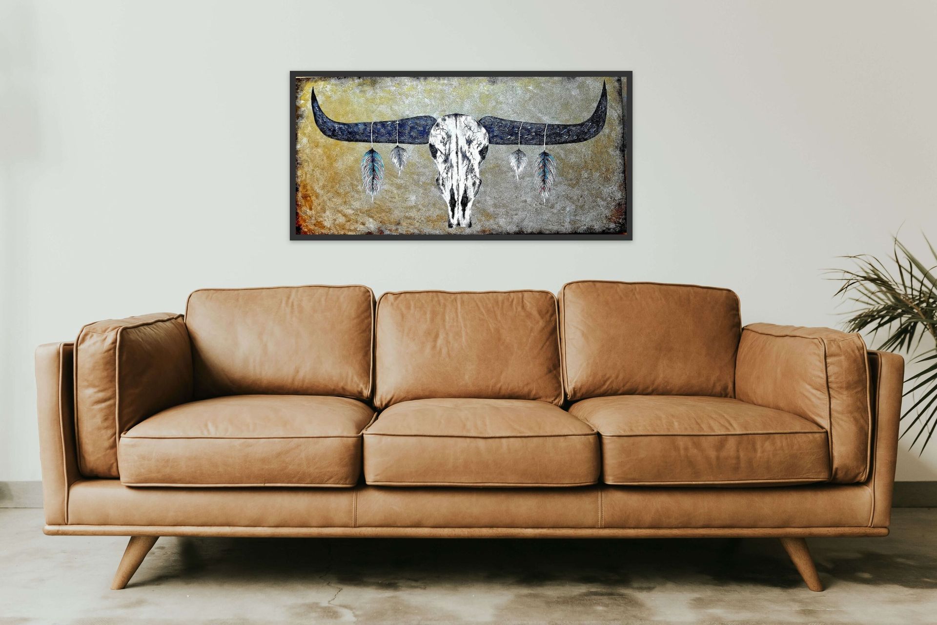 A longhorn cow skull artwork hangs on a light wall above a tan leather couch in a minimalist living room.