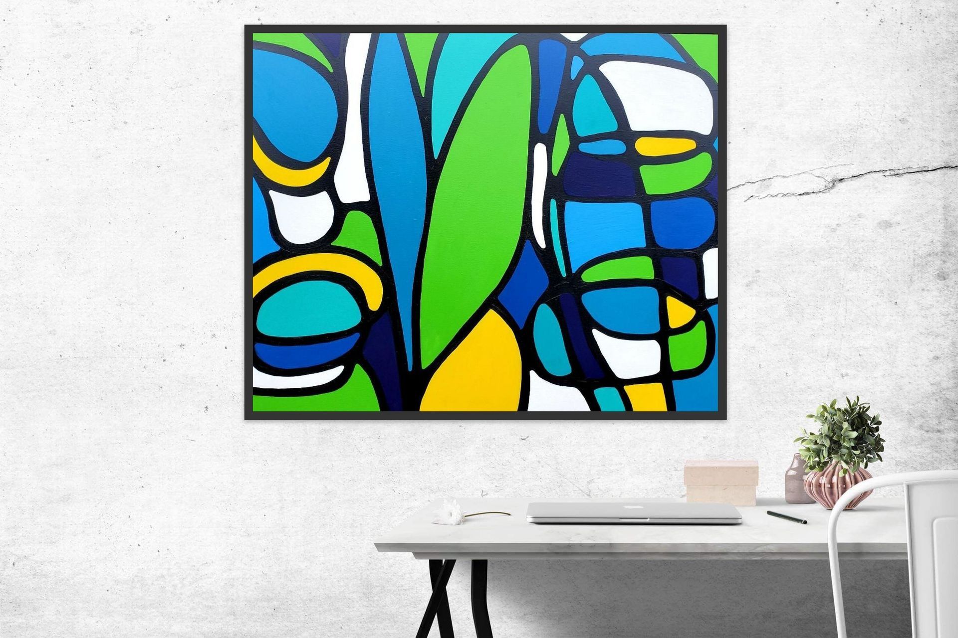 A framed abstract painting with blue, green, and yellow organic shapes hangs on a white wall above a desk and plant.