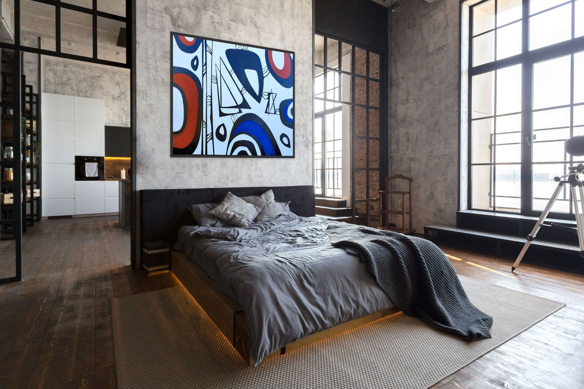 A modern, industrial-style bedroom featuring a grey bed, concrete walls, large windows, and abstract art above the bed.
