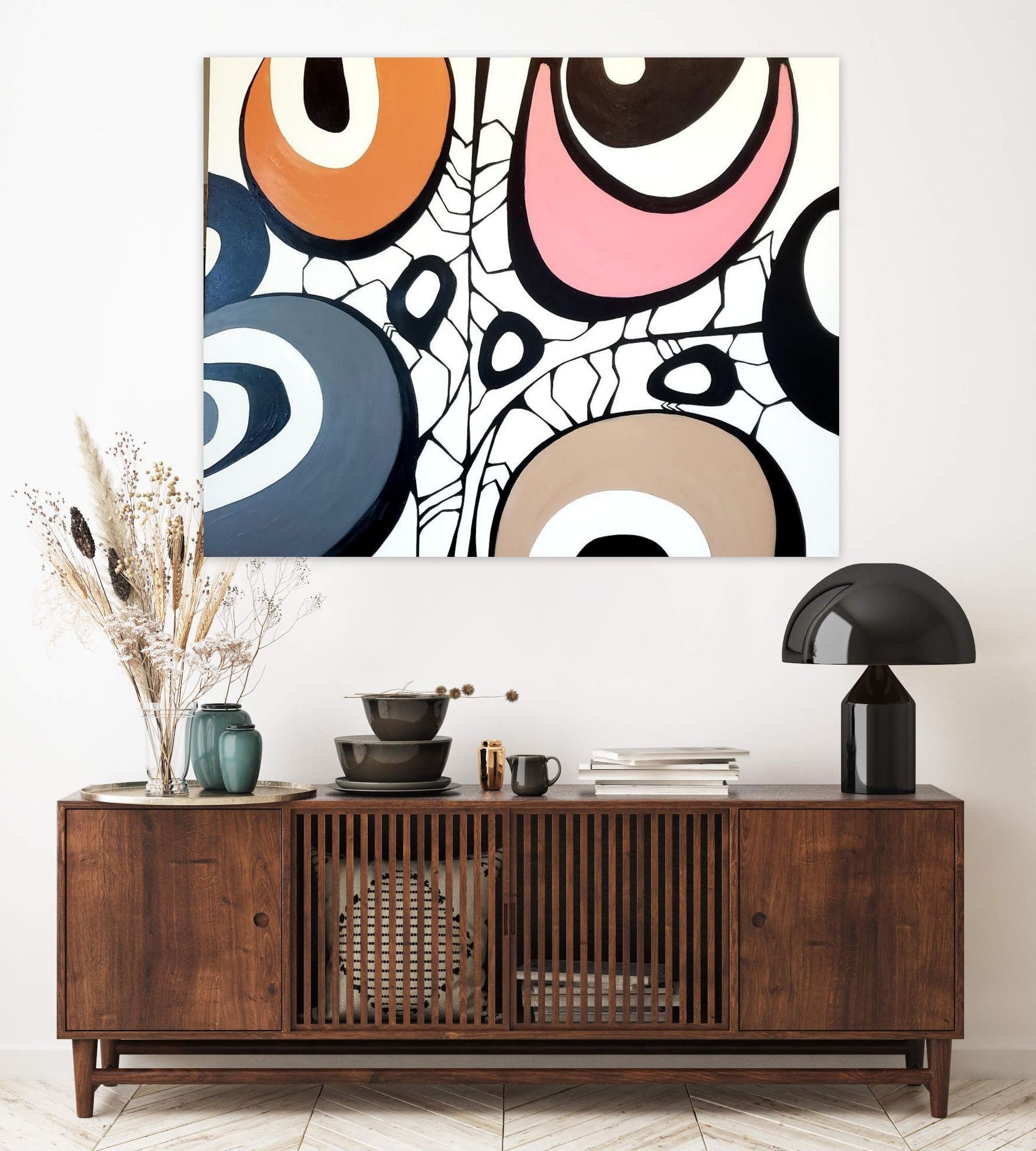 Abstract art with colorful circles hangs above a wooden mid-century sideboard with a dried floral arrangement and lamp.
