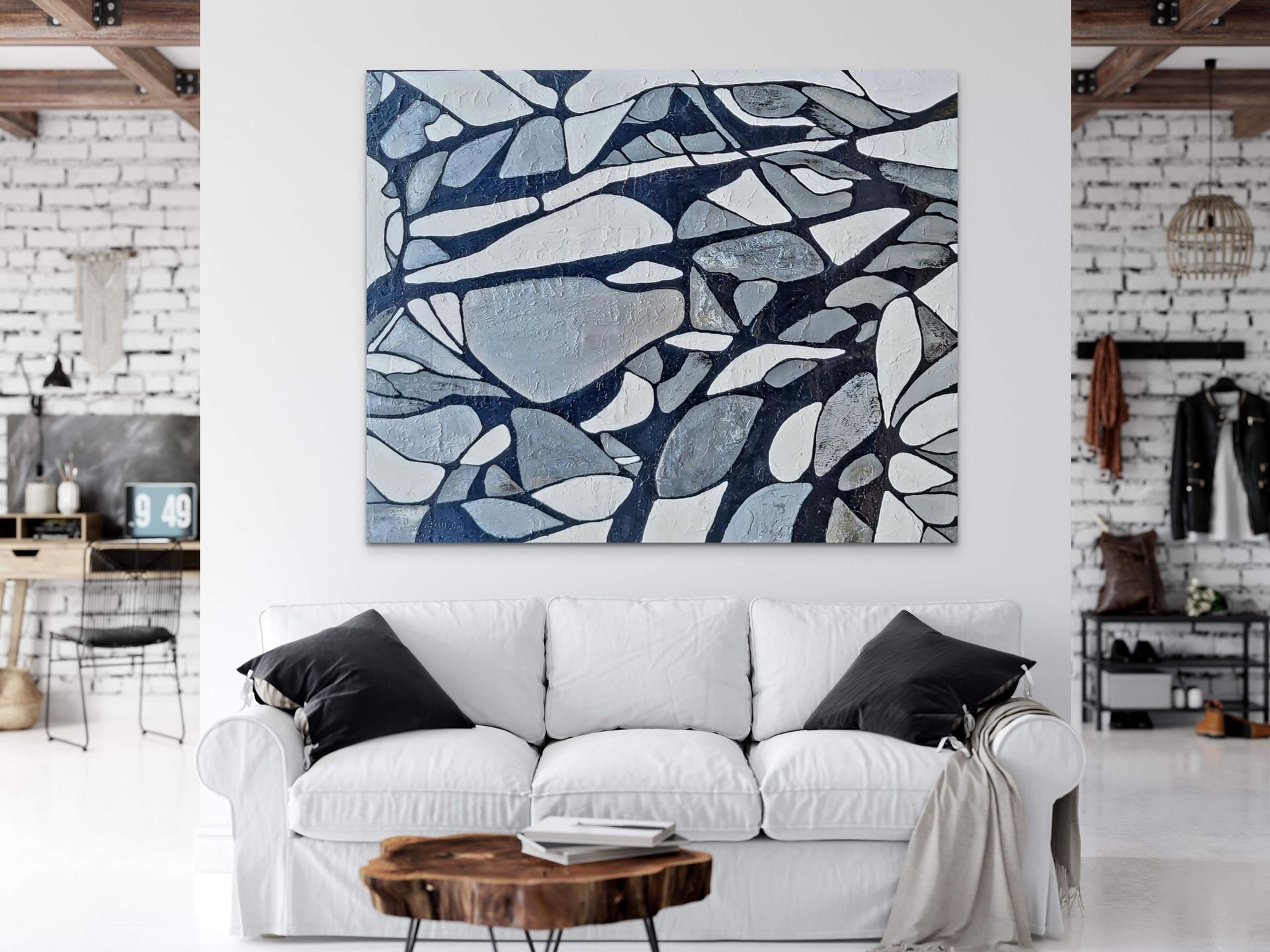 A white sofa with dark pillows sits under a large abstract painting with blue, grey, and white shapes in a bright room.