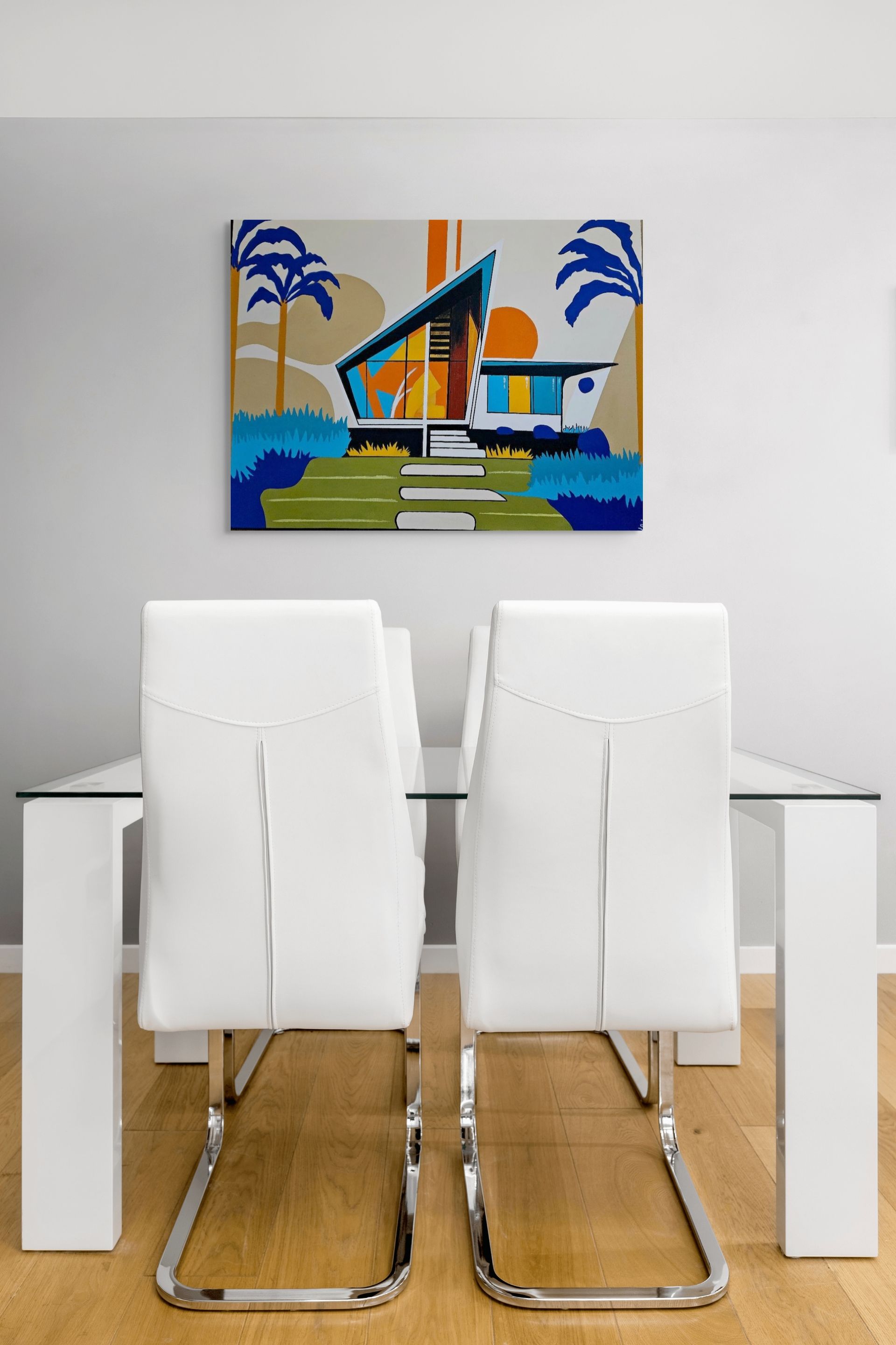 A mid-century modern style art print hangs above a wooden console table next to a white chair with a patterned pillow.