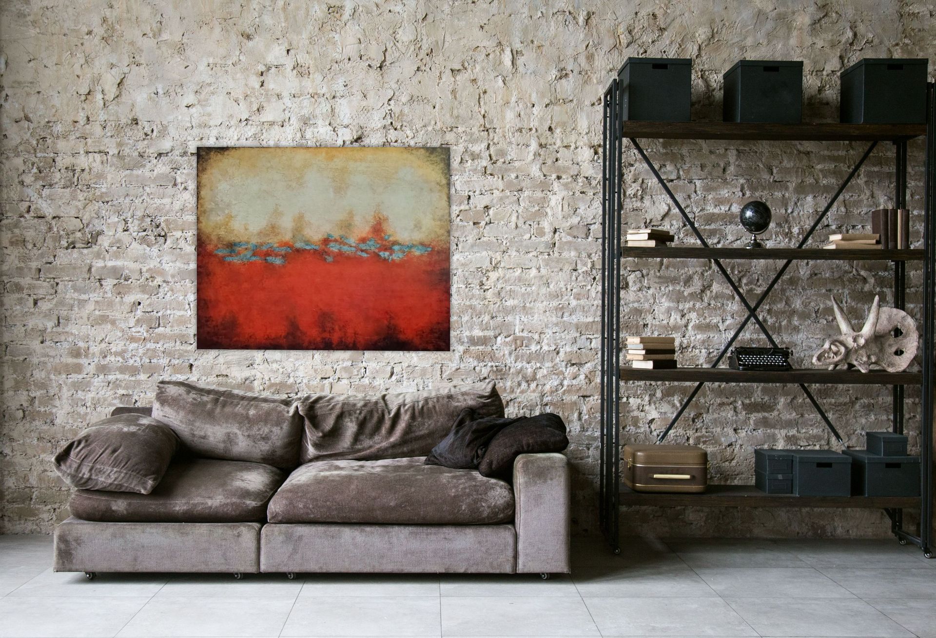 A brown sofa sits before a textured brick wall with abstract red and cream art above it and a metal bookshelf to the right.