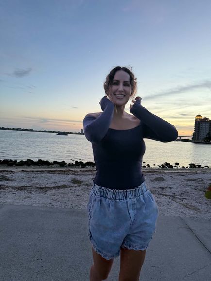 Person smiling on a beach at sunset, holding their face with the sea and sky behind them