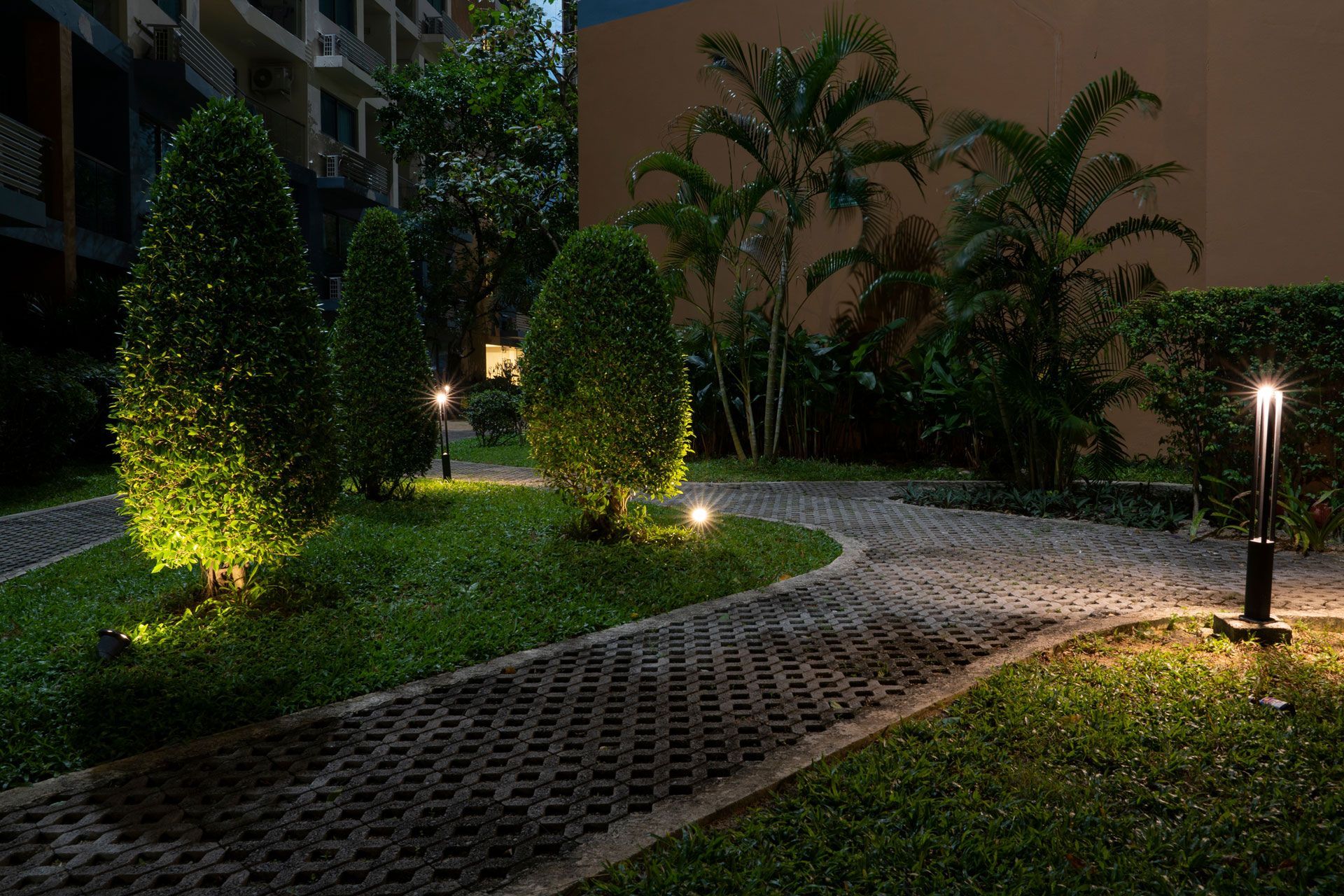 A illuminated paved garden pathway surrounded by manicured shrubs, palm trees, and soft landscape lighting at dusk.