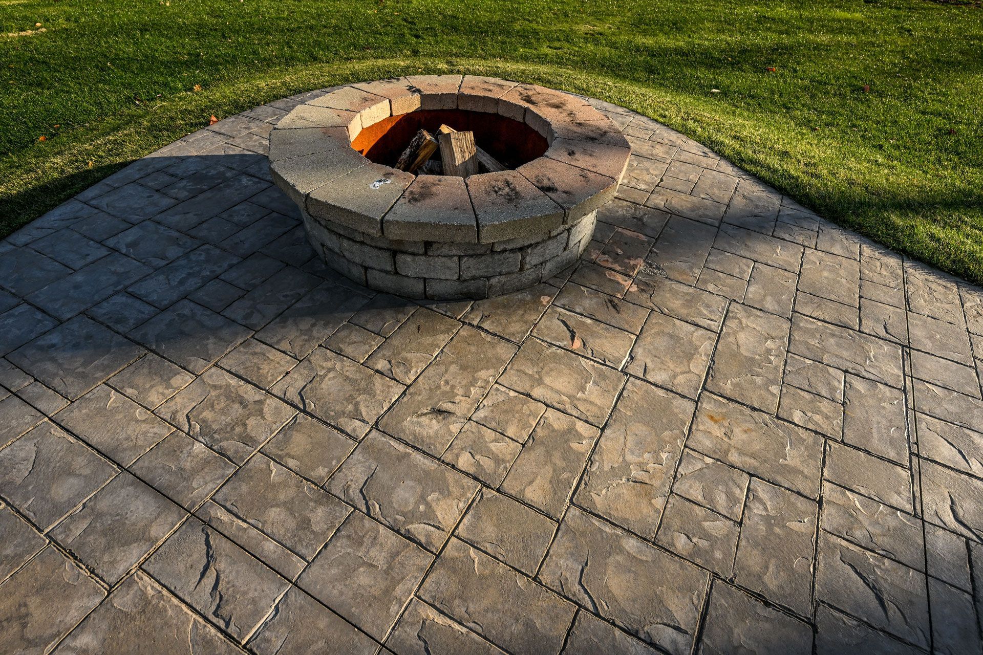 A circular stone fire pit sits on a gray patterned patio stone surface, surrounded by green grass.