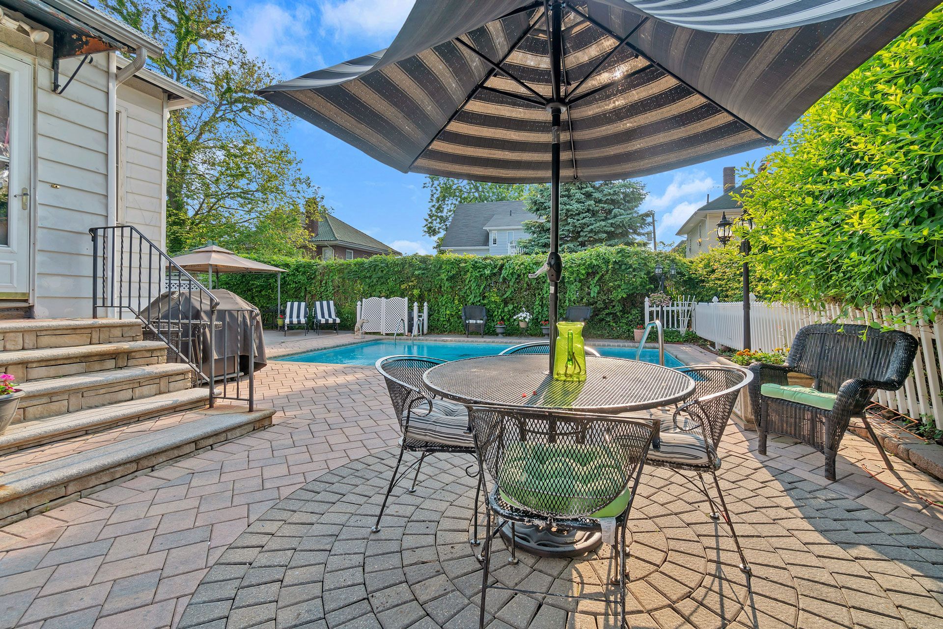 A patio with a round metal table, chairs, and an umbrella, featuring a pool and hedges in the background.