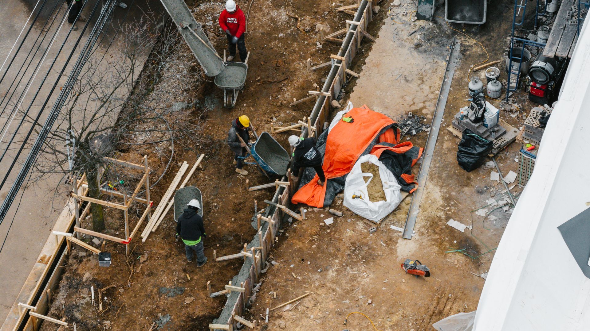 Aerial view of construction workers in hard hats pouring concrete and working near a fenced trench at a site.