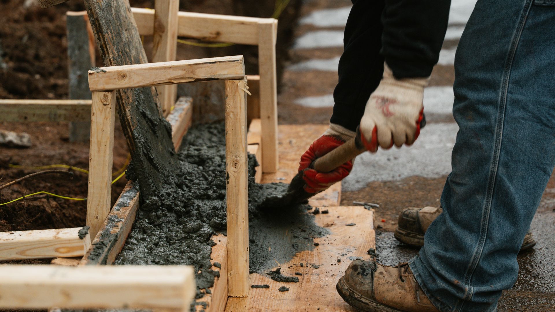 A person in work gloves uses a shovel to move wet concrete into a wooden frame at a construction site.
