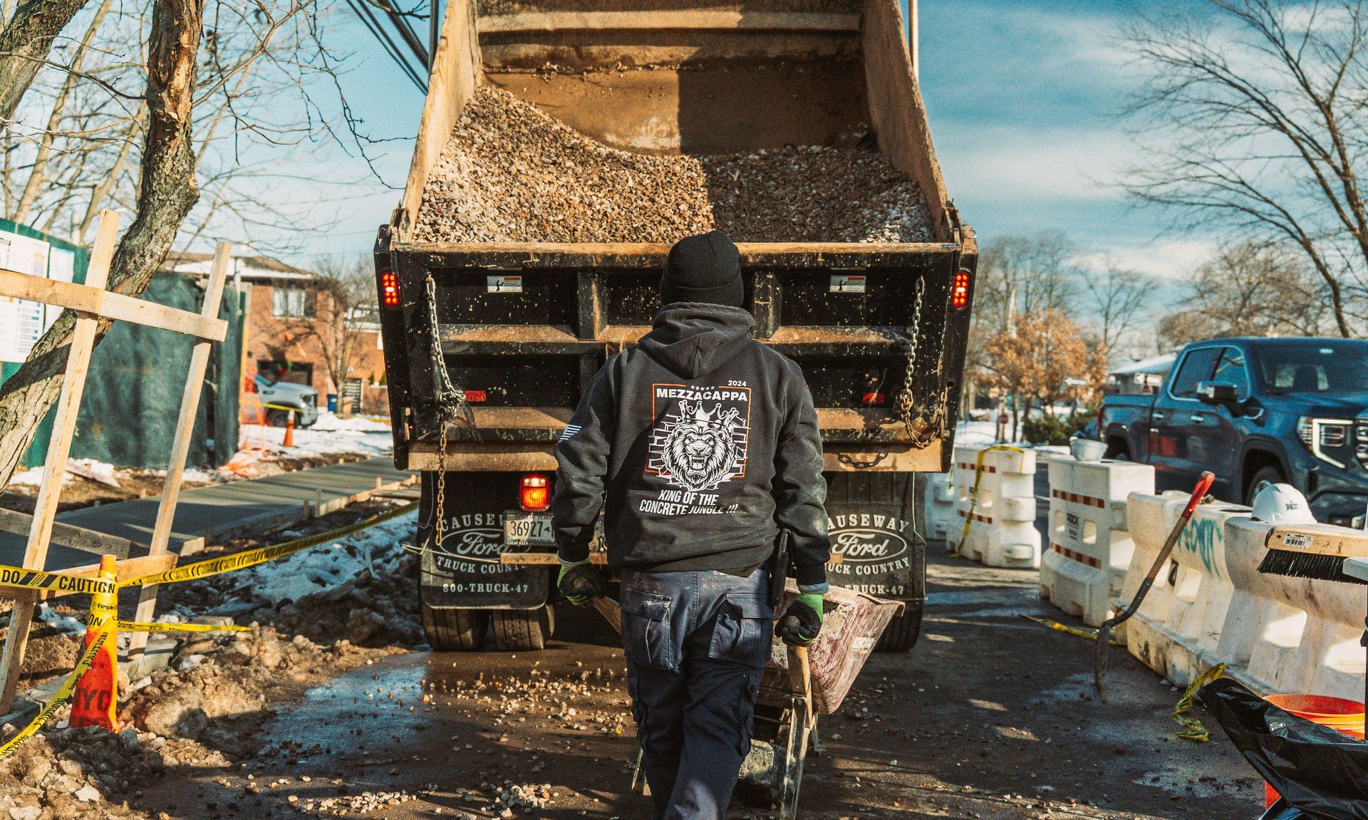 A person in a dark hoodie and work pants walks toward a dump truck filled with gravel at a construction site.