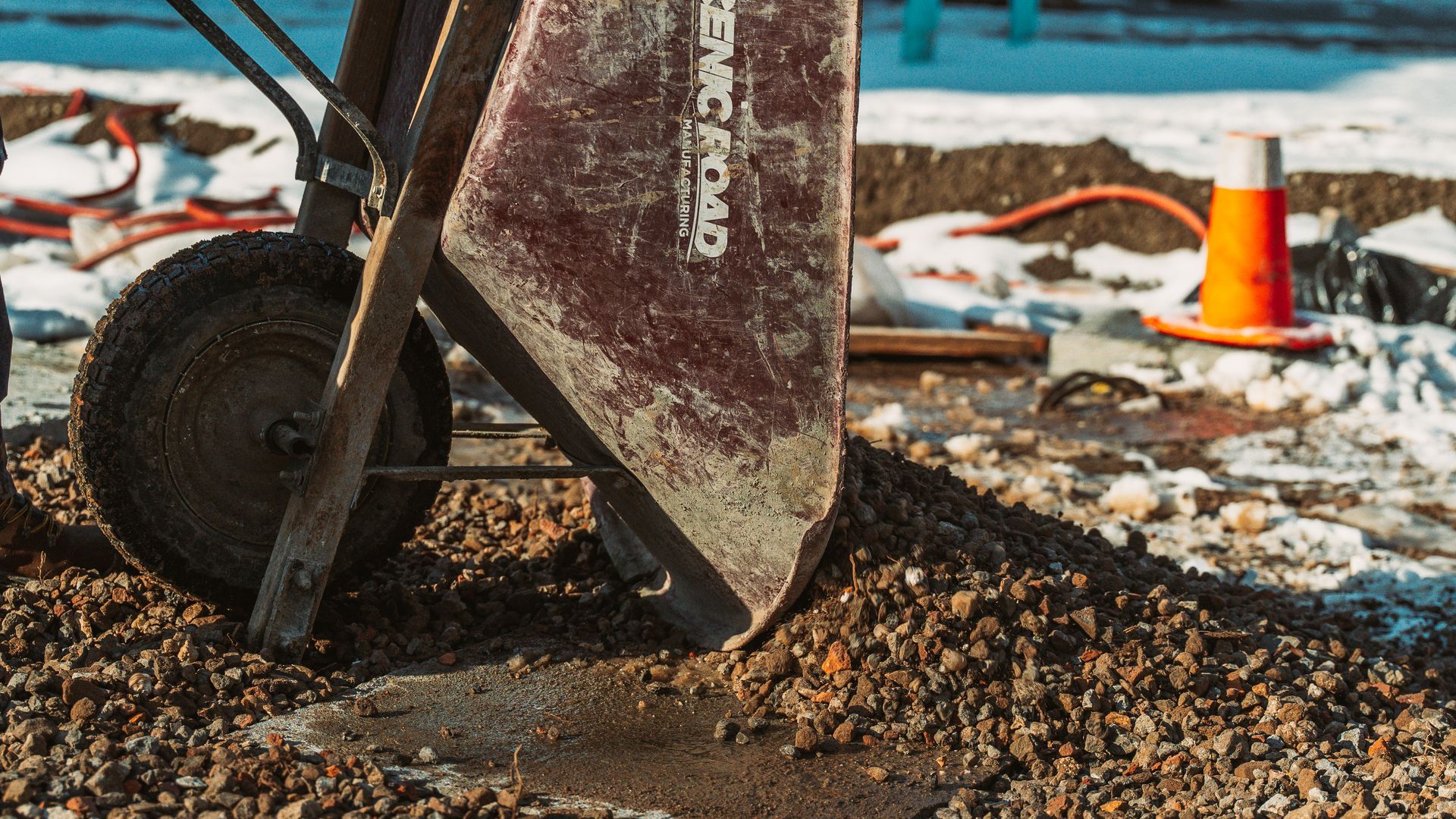 A worn wheelbarrow dumping gravel on a construction site, with a traffic cone in the snow-covered background.