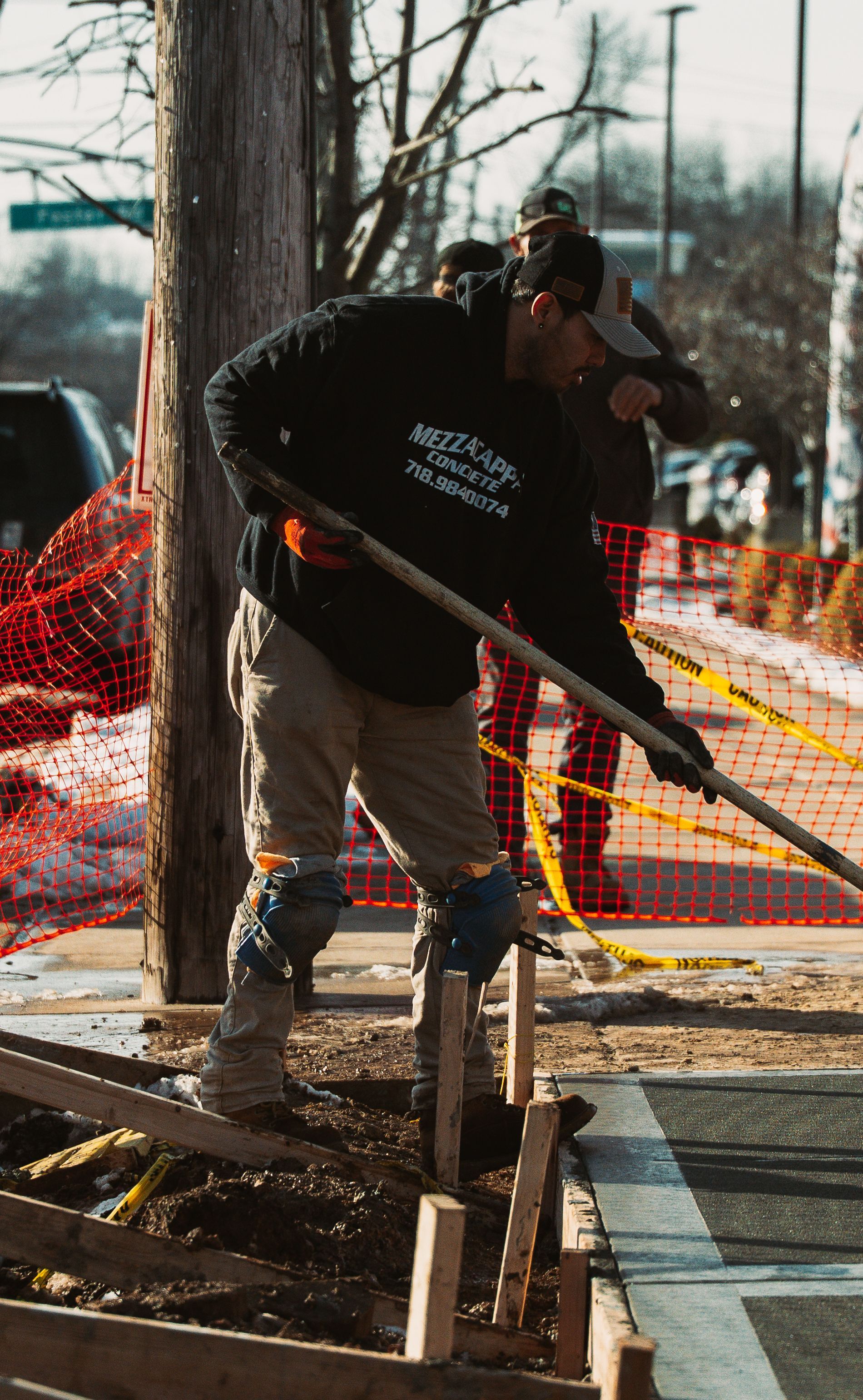 A construction worker in a black hoodie and knee pads uses a shovel to clear dirt near a wooden frame outdoors.