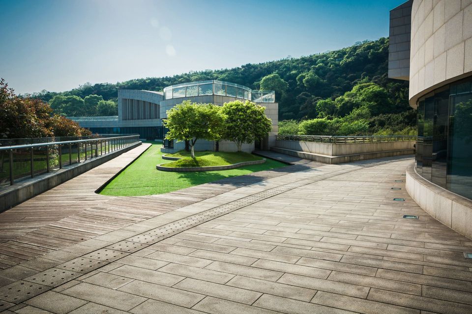 Modern outdoor terrace featuring a patch of lawn, two trees, and light stone paving against a backdrop of green hills.