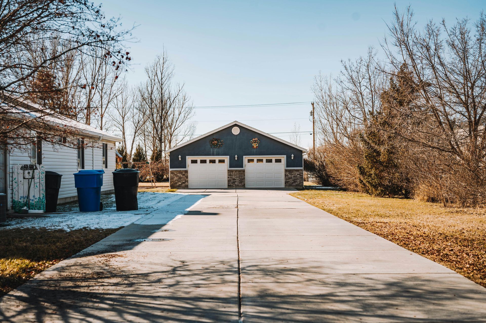 A concrete driveway leads to a gray two-car garage under a clear blue sky, with two trash bins outside a side building.