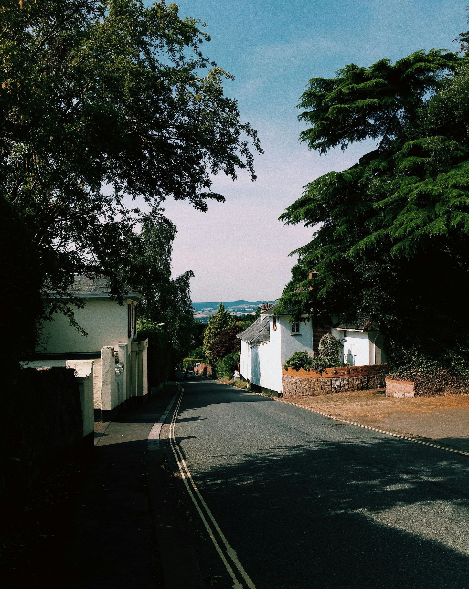 A tree-lined narrow road leads downhill past white cottages toward a distant view under a bright blue sky.