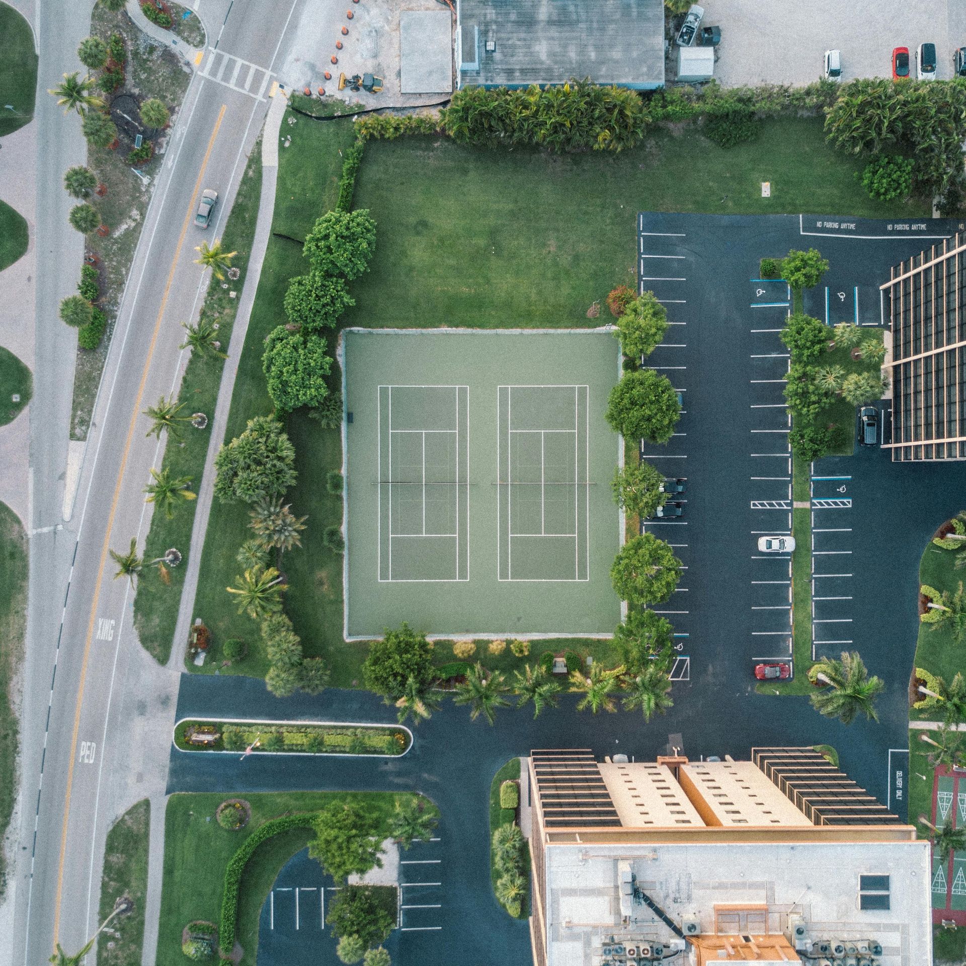 Aerial view of two green tennis courts situated in a parking lot next to a building and a road.