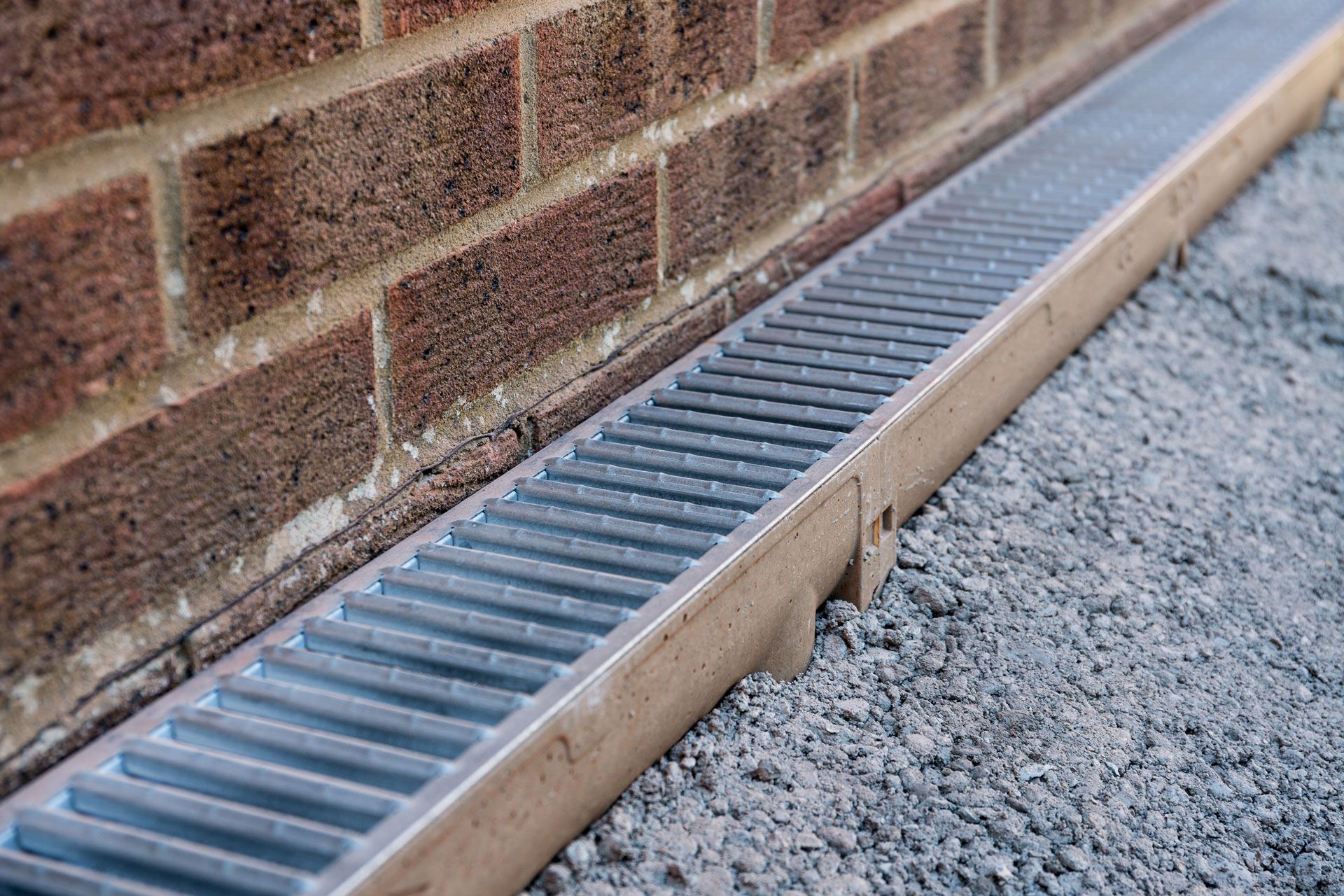 A metal channel drain installed along the base of a brick wall next to a gravel path.