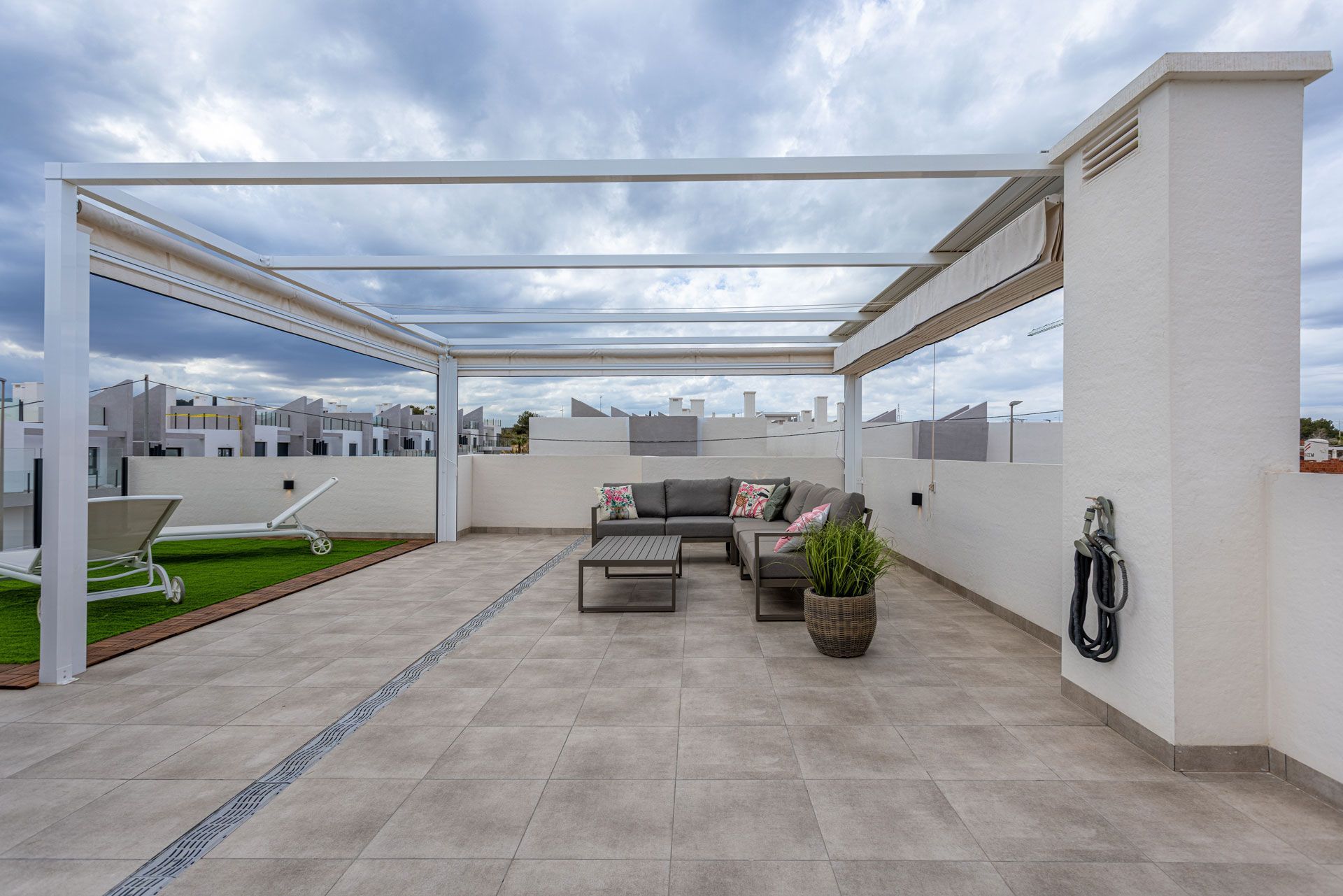 A rooftop terrace with tile flooring, a patio set under a white pergola, artificial grass, and a hose on a wall pillar.