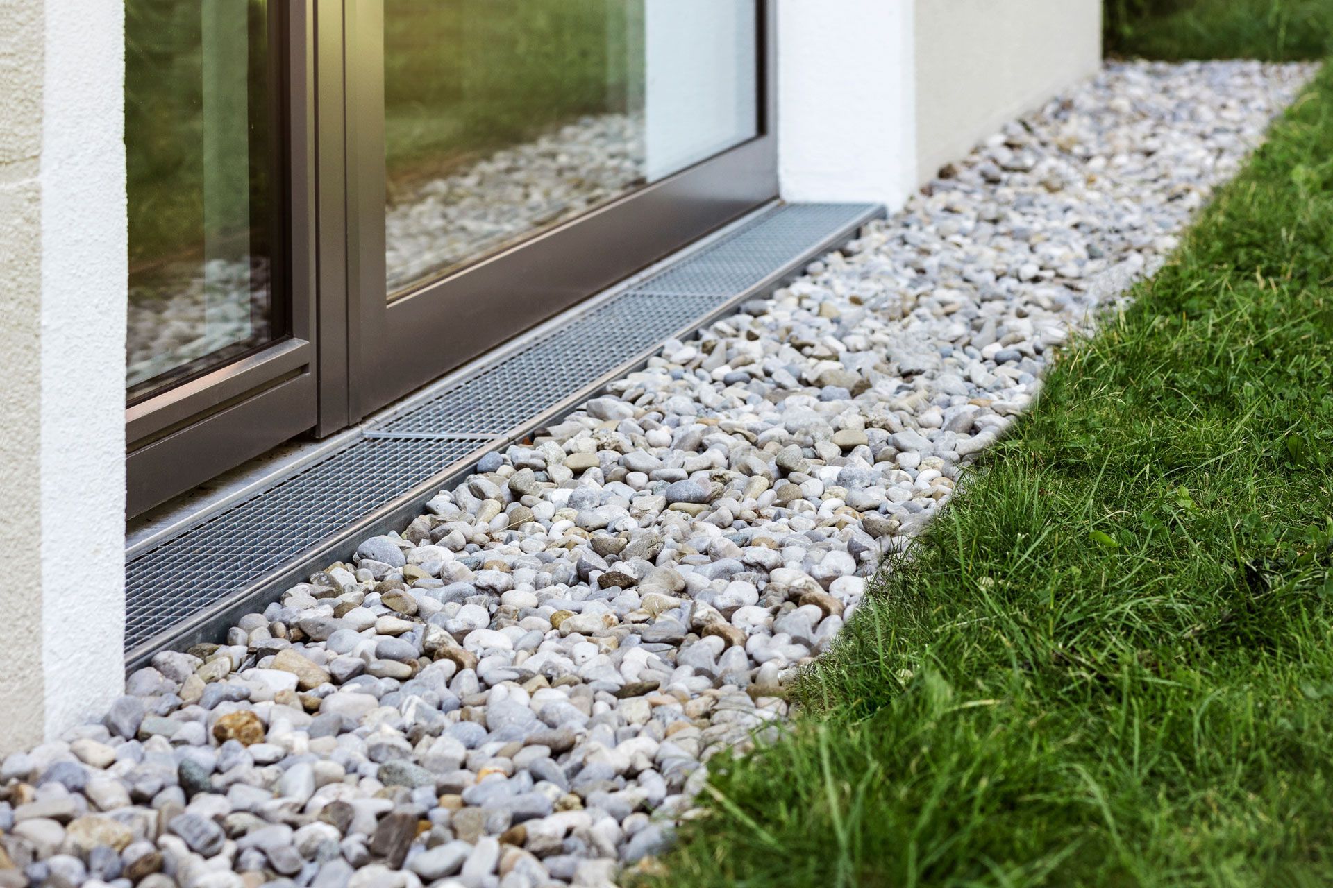 A metal drainage grate sits along the base of a glass patio door, surrounded by a border of grey landscaping gravel.
