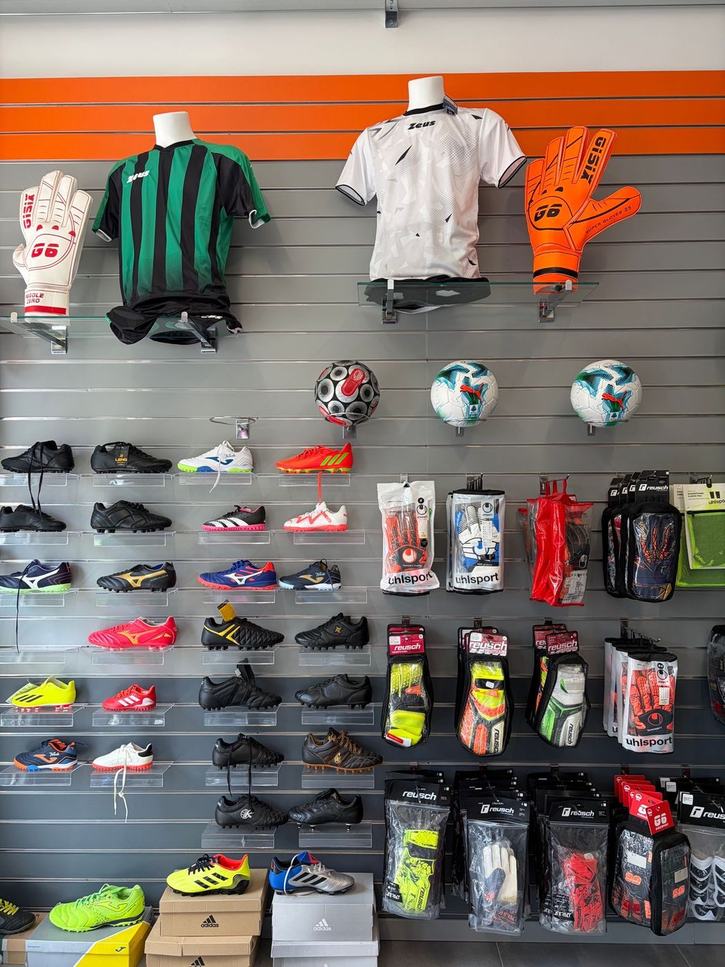 Espositore per abbigliamento sportivo con maglie, scarpe da calcio, parastinchi e palloni su mensole a doghe.