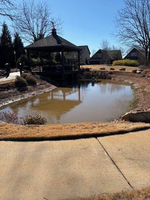 Pond beside a gazebo in a sunny landscaped park, with bare trees and a paved walkway nearby.