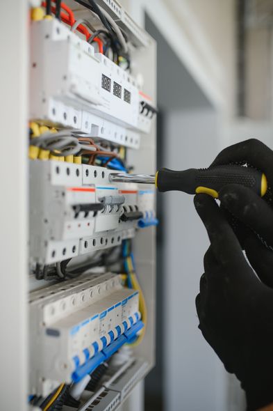 A person wearing black gloves uses a screwdriver to work on a wall-mounted electrical breaker panel with colorful wires.