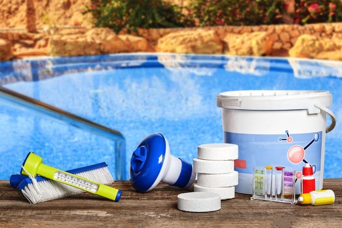 Poolside cleaning supplies beside a blue swimming pool, including brushes, bottles, buckets, and chemicals