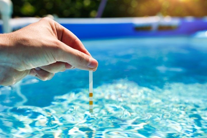 Hand holding a pool water test strip above a bright blue swimming pool