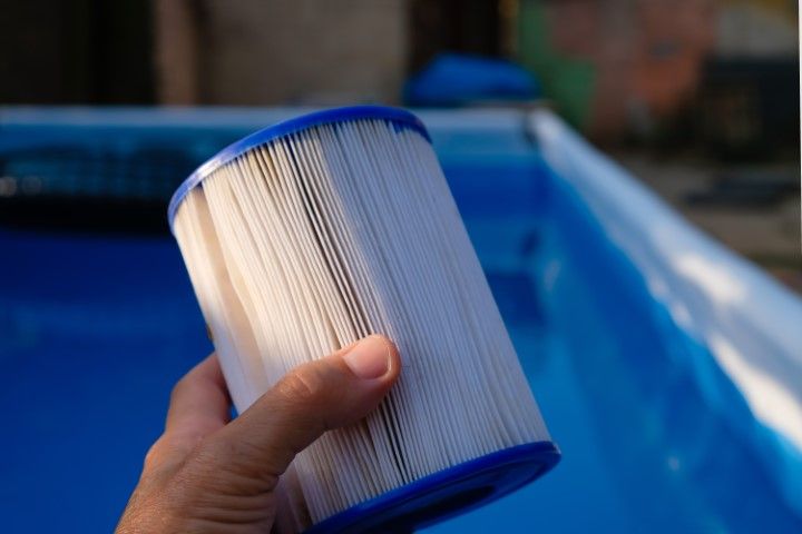 Hand holding a white and blue pool filter cartridge beside a blue swimming pool