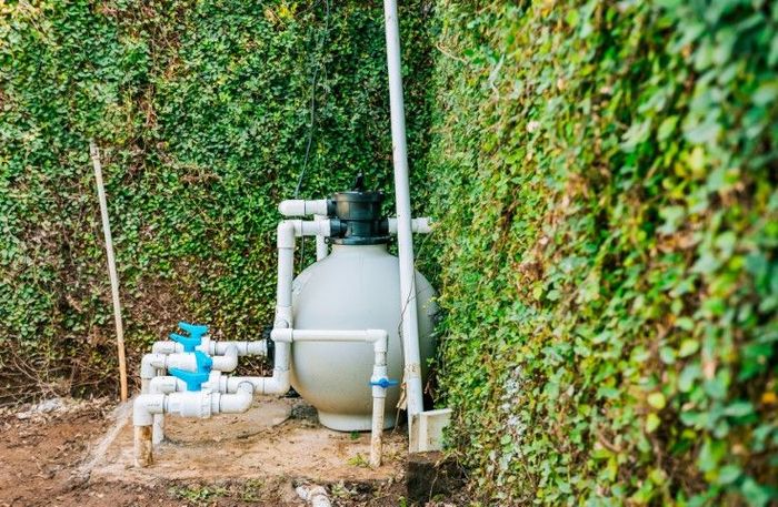 Water filtration tank and pipes beside a hedge-lined wall outdoors