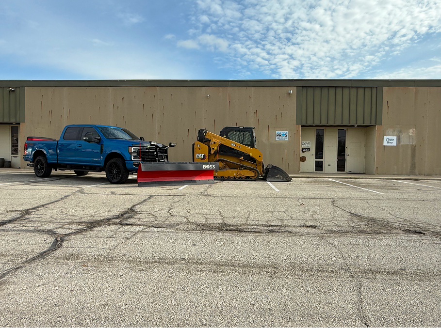 Blue truck with snowplow and yellow skid steer in front of a building.
