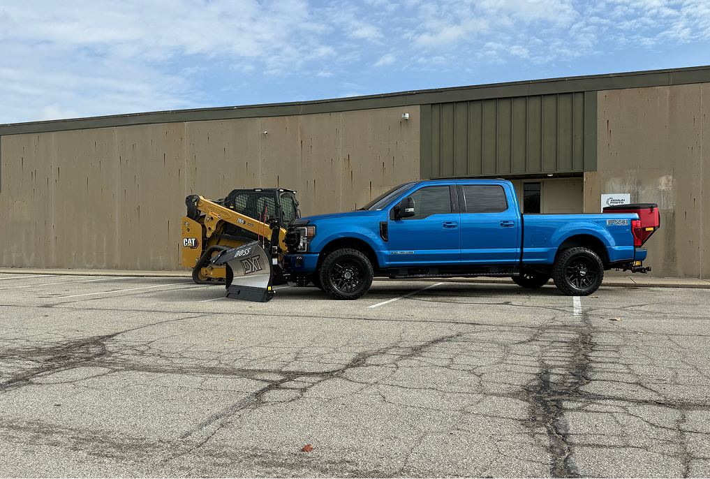 Blue pickup truck with snowplow attached, parked in front of a tan industrial building.