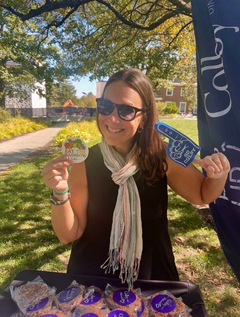 Woman at Colby College table, smiling, holding sticker and foam hand. Cookies on table, sunny outdoor setting.