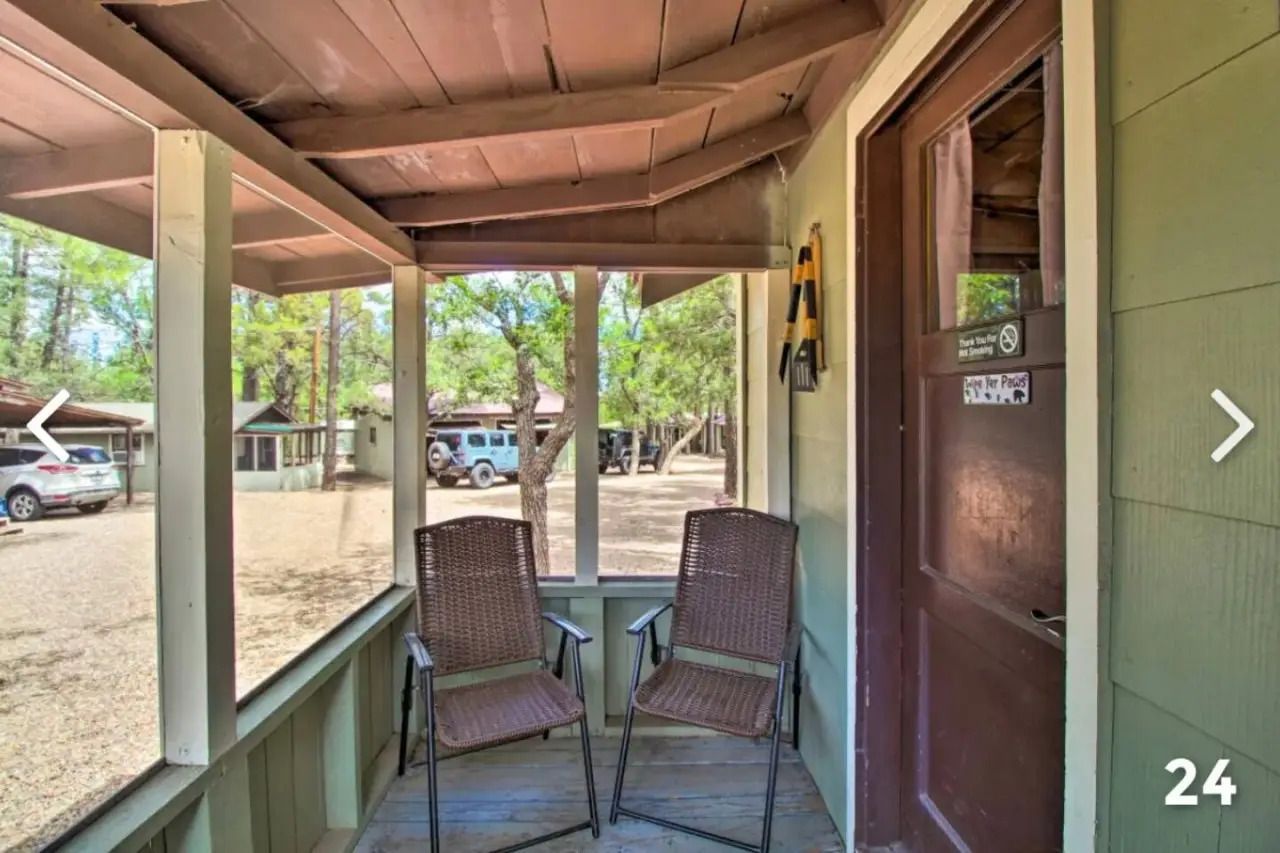 Covered front porch with two wicker chairs, a small table, and a brown door overlooking a gravel parking area.