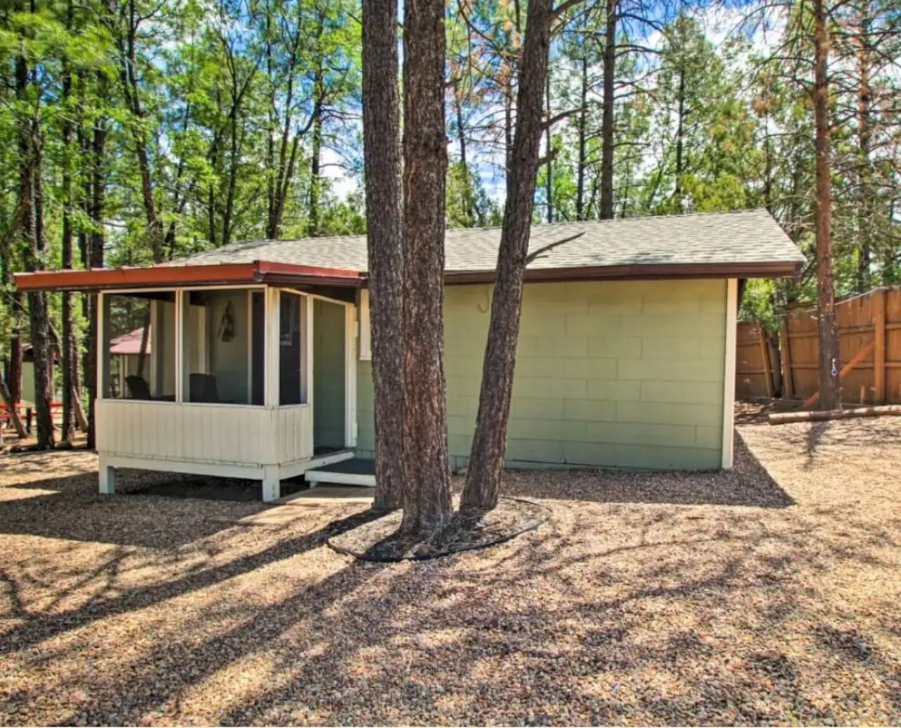 Small beige cabin with porch and red roof in a wooded yard