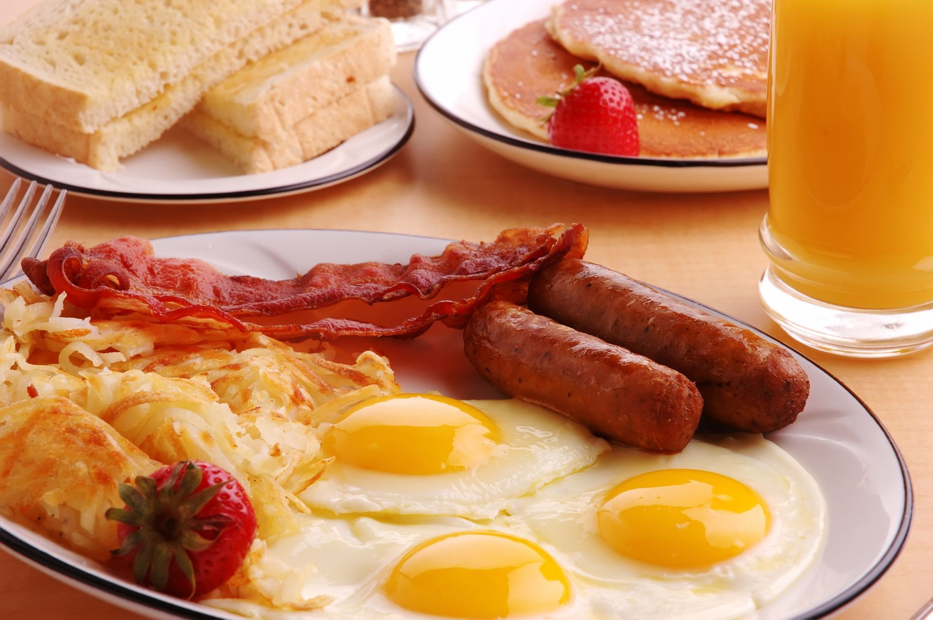Breakfast plate with fried eggs, bacon, sausage, hash browns, toast, pancakes, and orange juice