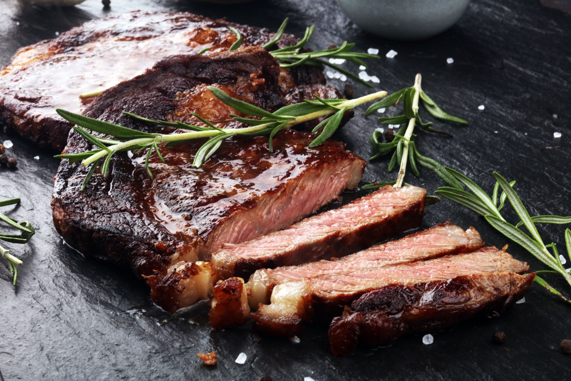 Sliced seared steak on a dark plate, garnished with rosemary and coarse salt.