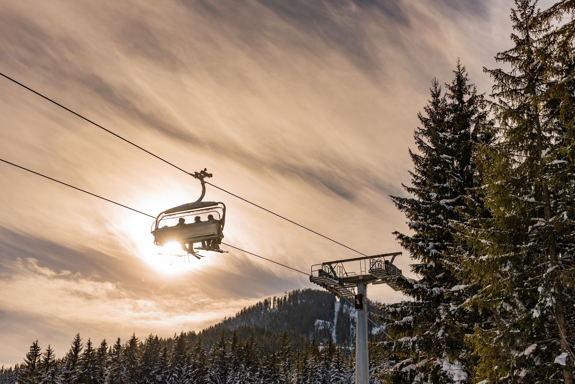 Chairlift gliding over snowy mountains at sunset, with pine trees silhouetted against a glowing sky