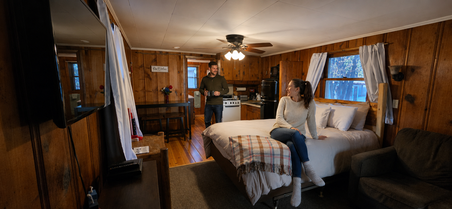 Cozy wood-paneled bedroom with a person sitting on a bed near a window and ceiling fan