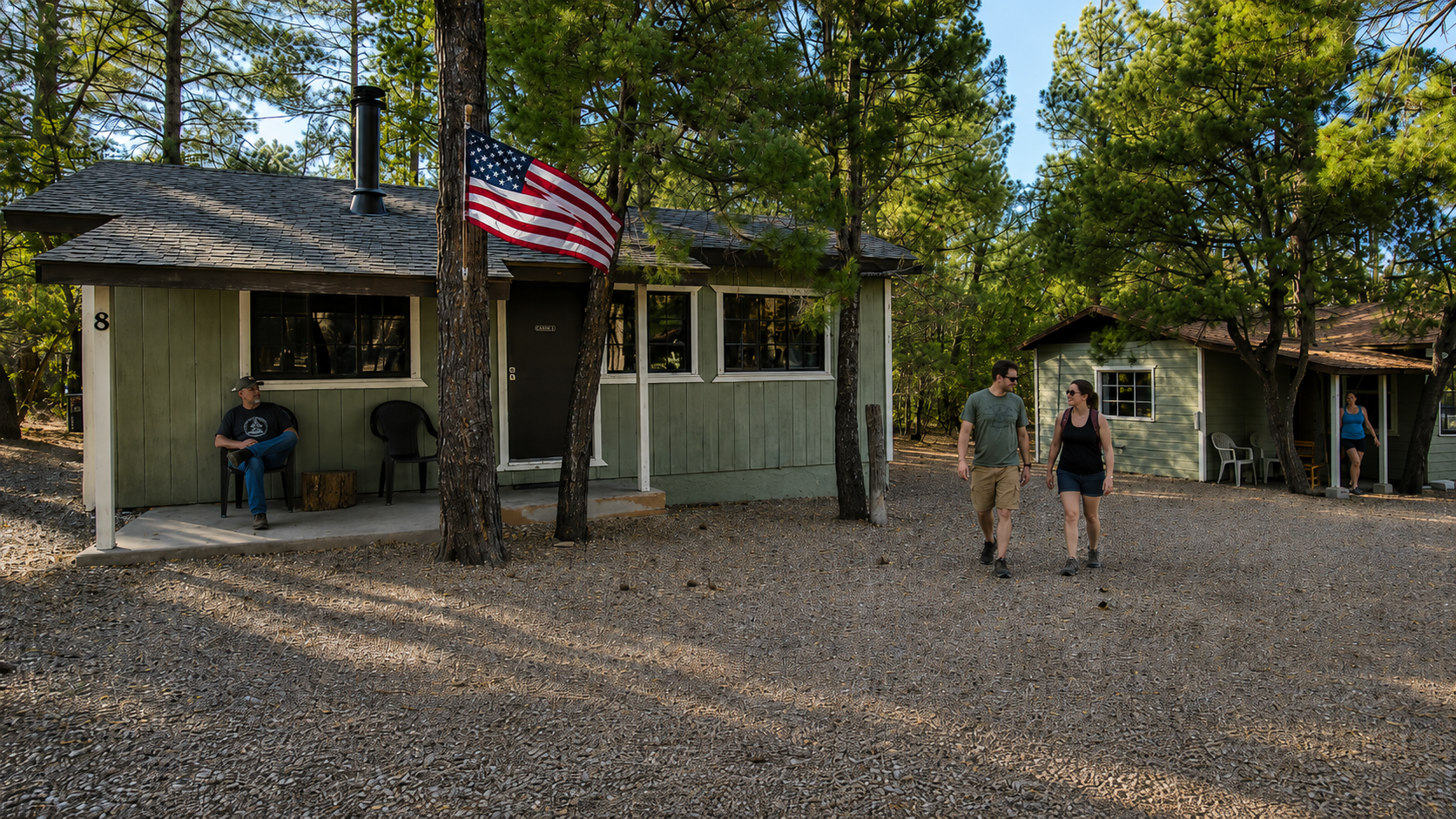 People standing outside rustic cabins in a wooded campground, with an American flag hanging in front.