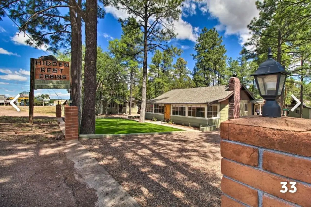 Wooded entrance with brick pillars, sign, lamp post, and a house under tall pine trees.