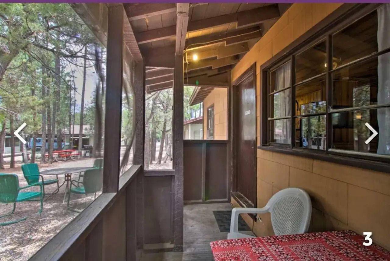 Covered cabin porch with white chairs, red tablecloth, and wooded outdoor seating area.