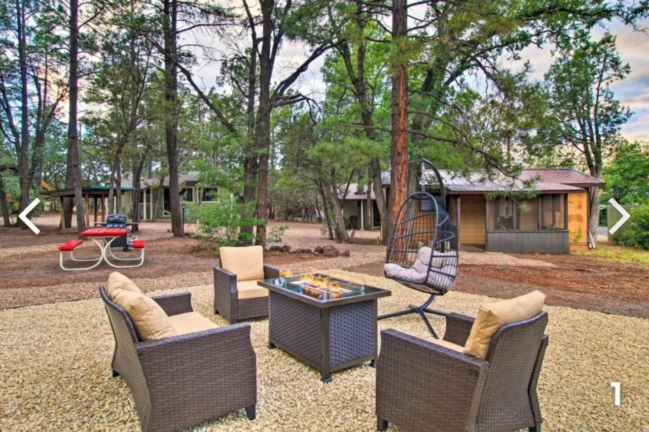Outdoor fire pit seating area with wicker chairs and a hanging swing under tall trees near cabins.