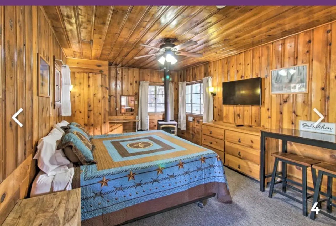 Rustic wood-paneled bedroom with quilted bed, dresser, wall TV, and desk under a ceiling fan.