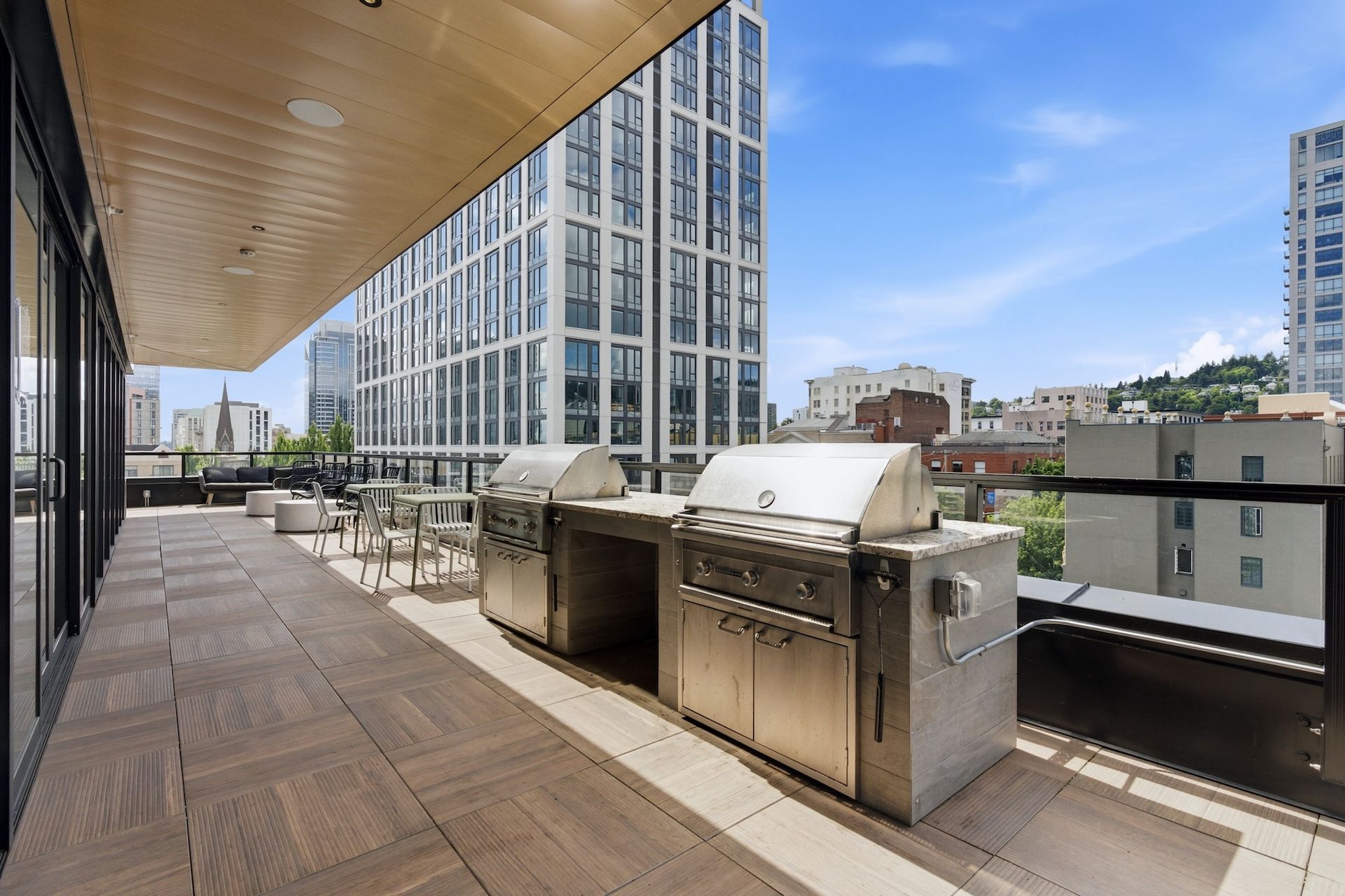 Rooftop terrace with stainless steel grills, wooden deck, and city buildings under a blue sky